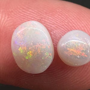 Puede incluir: Dos ópalos blancos con destellos iridiscentes de color. Uno es de forma ovalada, el otro es redondo. Los ópalos muestran un juego de colores con tonos amarillos, naranjas y verdes. Las piedras preciosas están sobre un fondo color piel.