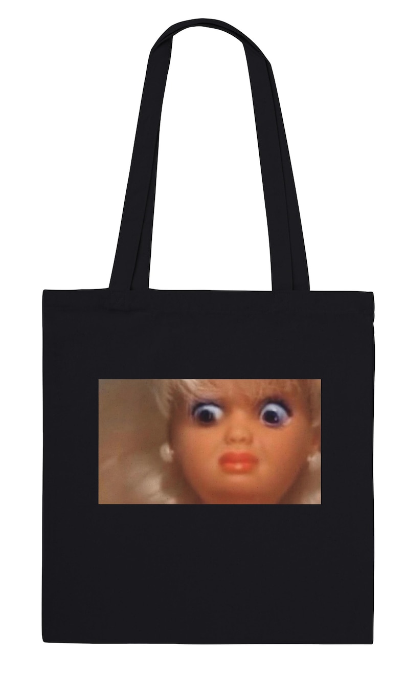 May include: Black tote bag with a close-up of a doll's face, showing wide eyes and a surprised expression. The doll has blonde hair, blue eyes, and red lips. The bag has two black handles.