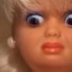 May include: Close-up of a doll's face with wide, blue eyes and a surprised expression. The doll has blonde hair, red lips, and small white earrings. The image is brightly lit.