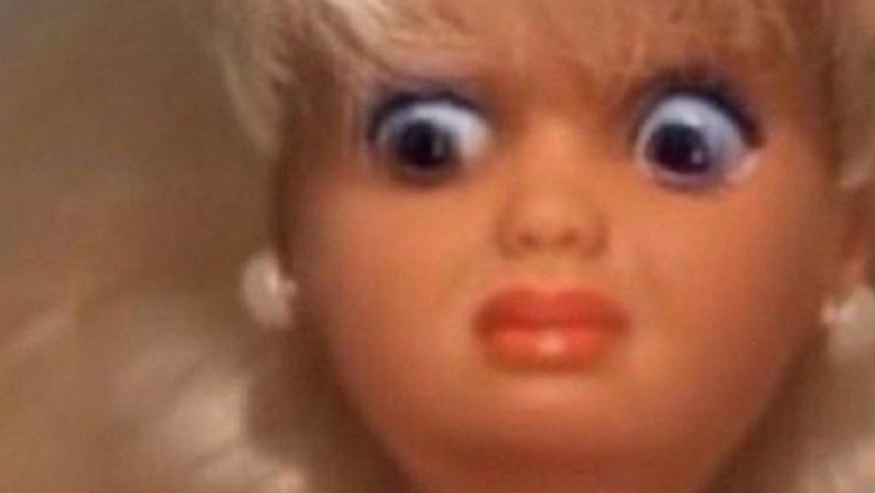May include: Close-up of a doll's face with wide, blue eyes and a surprised expression. The doll has blonde hair, red lips, and small white earrings. The image is brightly lit.