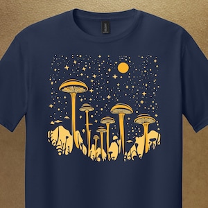 May include: A navy blue t-shirt with a yellow graphic depicting mushrooms under a starry night sky and a yellow moon. The design features detailed mushroom silhouettes and a celestial background, creating a whimsical and nature-inspired aesthetic.