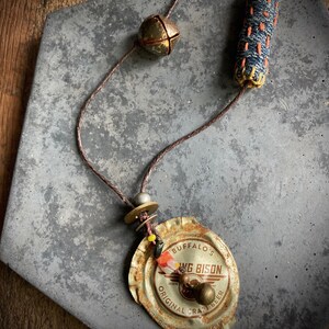 May include: A brown leather cord necklace with a rusty metal bottle cap pendant. The bottle cap has the words "Buffalo's Wing Bison Original Craft Beer" printed on it. The necklace also features a small bell, a blue and orange fabric charm, and a few beads.