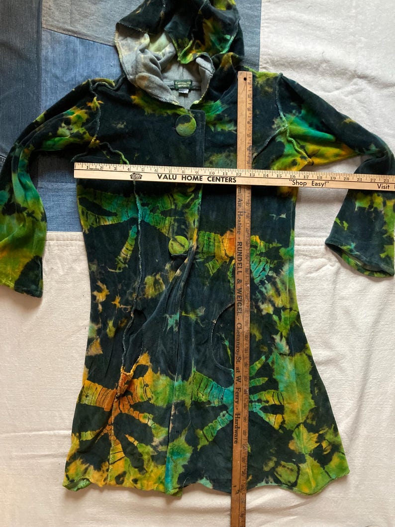 K&ouml;nnte beinhalten: Eine lange Kapuzenjacke mit Batikmuster in Gr&uuml;n-, Gelb- und Schwarzt&ouml;nen. Die Jacke hat einen Knopfverschluss und lange &Auml;rmel. Ein Holzlineal liegt auf der Jacke und misst ca. 76 cm. Der Text "VALU HOME CENTERS" ist sichtbar.