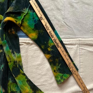 K&ouml;nnte beinhalten: Eine Jacke mit Batikmuster in Gr&uuml;n-, Gelb- und Blaut&ouml;nen. Die Jacke hat einen Knopfverschluss und lange &Auml;rmel. Ein Holzlineal liegt auf dem &Auml;rmel und misst ca. 63,5 cm.