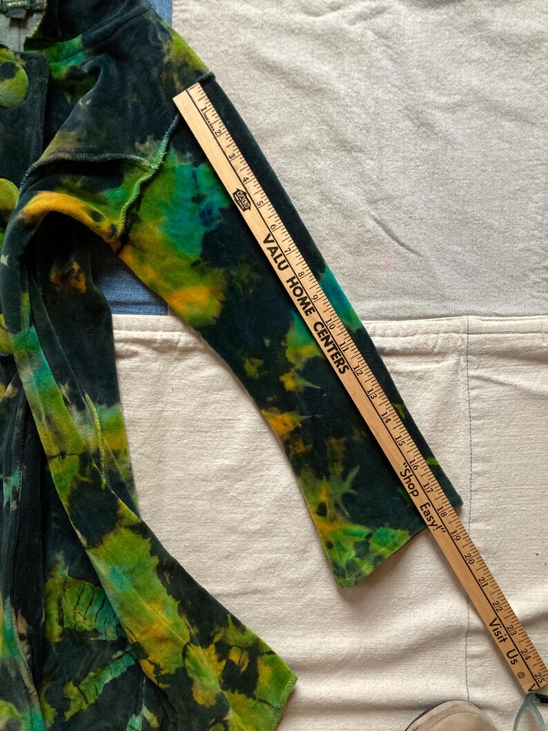 K&ouml;nnte beinhalten: Eine Jacke mit Batikmuster in Gr&uuml;n-, Gelb- und Blaut&ouml;nen. Die Jacke hat einen Knopfverschluss und lange &Auml;rmel. Ein Holzlineal liegt auf dem &Auml;rmel und misst ca. 63,5 cm.