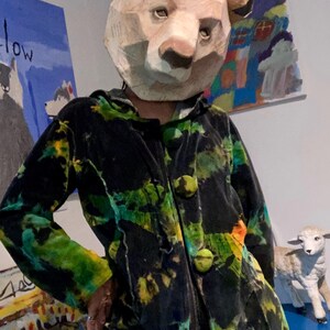 K&ouml;nnte beinhalten: Eine Person tr&auml;gt eine B&auml;renmaske aus Pappmach&eacute; und eine schwarze Jacke mit gr&uuml;n-gelben Batikmustern. Die Jacke hat gro&szlig;e, runde, gr&uuml;ne Kn&ouml;pfe. Kunstwerke sind im Hintergrund zu sehen.