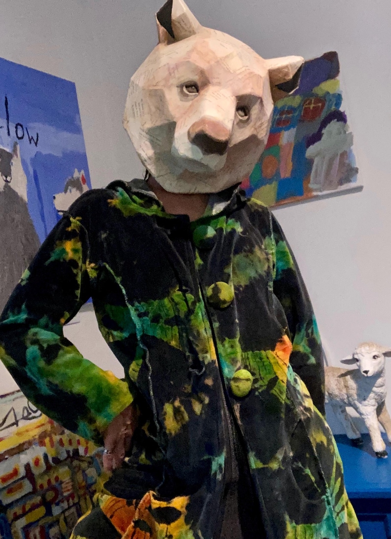 K&ouml;nnte beinhalten: Eine Person tr&auml;gt eine B&auml;renmaske aus Pappmach&eacute; und eine schwarze Jacke mit gr&uuml;n-gelben Batikmustern. Die Jacke hat gro&szlig;e, runde, gr&uuml;ne Kn&ouml;pfe. Kunstwerke sind im Hintergrund zu sehen.