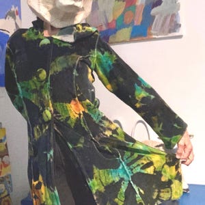 Puede incluir: Un abrigo largo negro con un vibrante estampado tie-dye en tonos verdes, amarillos y azules. El abrigo presenta botones grandes y redondos y un diseño artístico único. La persona lleva una máscara de oso de papel maché.