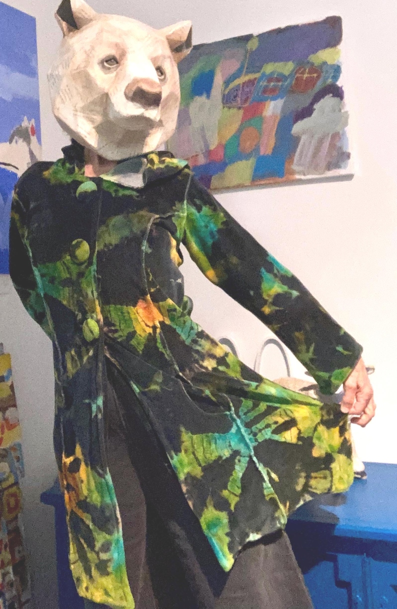 K&ouml;nnte beinhalten: Ein langer, schwarzer Mantel mit einem lebendigen Batikmuster in Gr&uuml;n-, Gelb- und Blaut&ouml;nen. Der Mantel hat gro&szlig;e, runde Kn&ouml;pfe und ein einzigartiges, k&uuml;nstlerisches Design. Die Person tr&auml;gt eine Pappmach&eacute;-B&auml;renmaske.