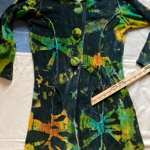 K&ouml;nnte beinhalten: Eine lang&auml;rmlige Jacke mit Batikmuster und Kapuze. Die Jacke hat eine dunkle Grundfarbe mit leuchtenden gr&uuml;nen, gelben und orangenen Mustern. Sie hat drei gro&szlig;e, runde Kn&ouml;pfe auf der Vorderseite und Ausschnitte. Der Stoff scheint ein weiches, samtartiges Material zu sein.