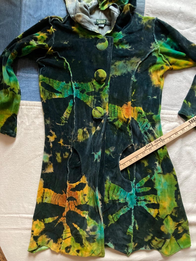 K&ouml;nnte beinhalten: Eine lang&auml;rmlige Jacke mit Batikmuster und Kapuze. Die Jacke hat eine dunkle Grundfarbe mit leuchtenden gr&uuml;nen, gelben und orangenen Mustern. Sie hat drei gro&szlig;e, runde Kn&ouml;pfe auf der Vorderseite und Ausschnitte. Der Stoff scheint ein weiches, samtartiges Material zu sein.