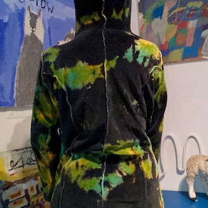 K&ouml;nnte beinhalten: Ein langer Kapuzenmantel mit einem Batikmuster in Schwarz, Gr&uuml;n und Gelb. Der Mantel scheint aus einem weichen Material zu sein. Die R&uuml;ckseite des Mantels ist sichtbar, mit einer Naht in der Mitte. Kunstwerke sind im Hintergrund zu sehen.