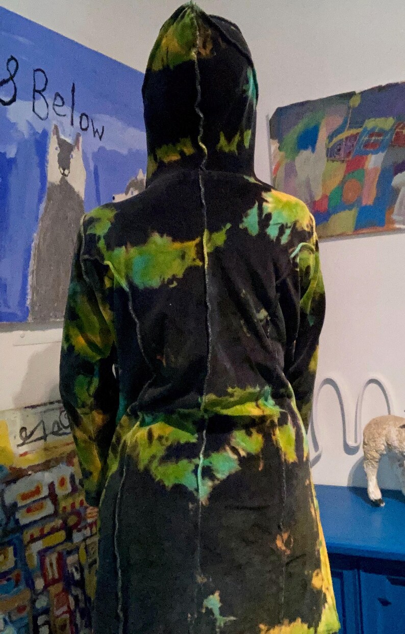 K&ouml;nnte beinhalten: Ein langer Kapuzenmantel mit einem Batikmuster in Schwarz, Gr&uuml;n und Gelb. Der Mantel scheint aus einem weichen Material zu sein. Die R&uuml;ckseite des Mantels ist sichtbar, mit einer Naht in der Mitte. Kunstwerke sind im Hintergrund zu sehen.