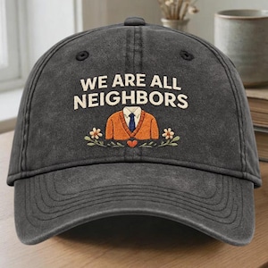 May include: A dark grey baseball cap with the text "WE ARE ALL NEIGHBORS" in white. The cap features an embroidered design of an orange sweater, a blue tie, and floral accents. The hat is made of a durable fabric.