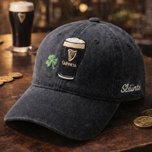 May include: A black denim baseball cap featuring an embroidered Guinness pint glass and a green shamrock. The word "Sl&aacute;inte" is embroidered on the side. A pint of Guinness is in the background.