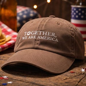 May include: A brown baseball cap with the embroidered text "TOGETHER, WE ARE AMERICA" in white. The cap has a curved peak and is made of a soft fabric. The background includes a red, white, and blue theme.