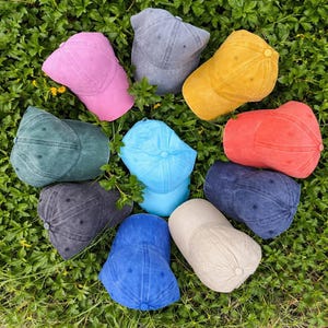 May include: A collection of ten colorful baseball caps in various shades, including pink, gray, yellow, red, blue, and green, arranged on a bed of green foliage. Each cap has a curved brim and a button on top.