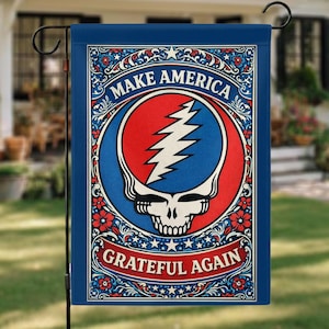 Op de afbeelding: Een blauwe tuin vlag met de tekst "MAKE AMERICA GRATEFUL AGAIN" in een decoratieve lijst. De vlag heeft een rood, wit en blauw ontwerp met een schedel en bliksem.