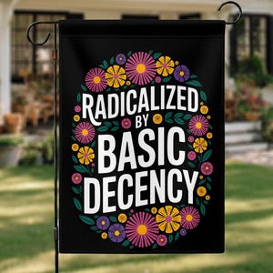 Op de afbeelding: Zwarte tuin vlag met de witte tekst "RADICALIZED BY BASIC DECENCY", omringd door een bloemenkrans van roze, gele en paarse bloemen met groene bladeren. De vlag hangt in een tuin.