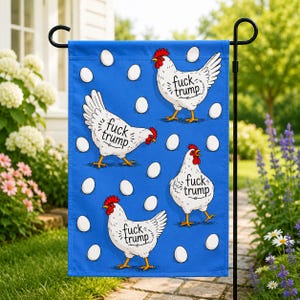 May include: A rectangular blue garden flag featuring a repeating pattern of white eggs and three white chickens with red combs and wattles. Each chicken has the text "fuck trump" printed in black on its side, displayed on a black metal stand.