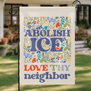 Op de afbeelding: Een tuinvaandel met de woorden "ABOLISH ICE" in blauw en "LOVE THY neighbor" in oranje en blauw. De vlag heeft een bloemenpatroon op de achtergrond met bloemen in verschillende kleuren, waaronder blauw, rood en geel.
