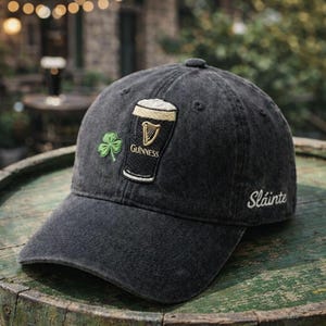 May include: A black baseball cap with a Guinness logo, a green shamrock, and the word "Sl&aacute;inte" embroidered in white. The cap has a curved brim and a button on top. The background is blurred, suggesting an outdoor setting.