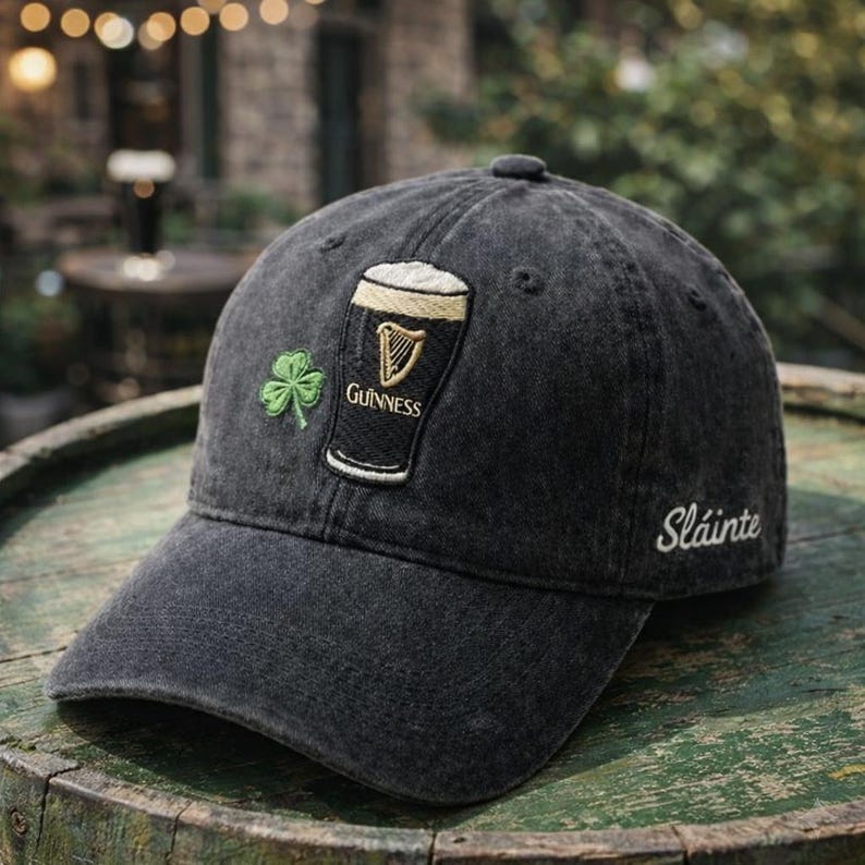May include: A black baseball cap with a Guinness logo, a green shamrock, and the word "Sl&aacute;inte" embroidered in white. The cap has a curved brim and a button on top. The background is blurred, suggesting an outdoor setting.