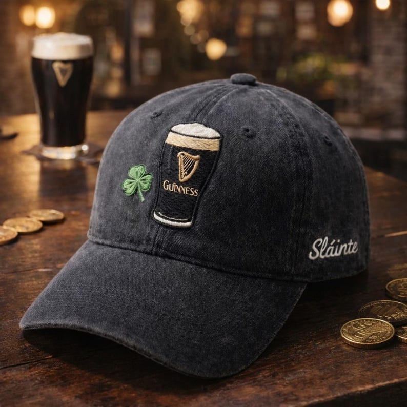 May include: A black denim baseball cap featuring an embroidered Guinness pint glass and a green shamrock. The word "Sl&aacute;inte" is embroidered on the side. A pint of Guinness is in the background.