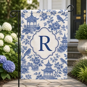 May include: A garden flag with a blue and white toile pattern, depicting pagodas, birds, and flowers. The flag has a white central panel with a large blue letter "R". It is displayed on a black metal stand.