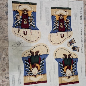 May include: A white fabric panel with four Christmas stocking designs. Each design features a figure with angel wings, birdhouses, and a snowy landscape. The text "WHISPERING PINES CHRISTMAS" is printed on the right side.