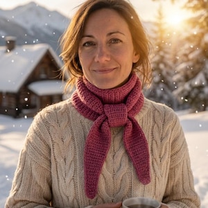 May include: A dusty rose crochet neck scarf tied in a simple knot, worn over a cream-colored cable knit sweater. The scarf features a textured, dense stitch pattern with tapered ends, providing a solid color contrast against the neutral knit.