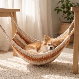 Op de afbeelding: Een kleine, langharige tan en witte hond slaapt in een gehaakte hangmat. De hangmat heeft horizontale strepen in tinten crème, beige en roestoranje, opgehangen aan dikke, gevlochten touwen bevestigd aan twee houten poten.