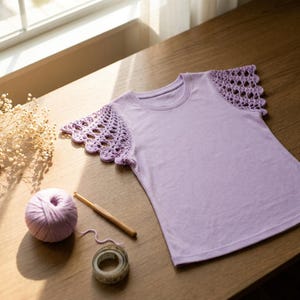 May include: A light purple t-shirt featuring short, flared sleeves made of intricate, openwork crochet lace. The garment has a round neckline and a smooth, solid-colored bodice. A ball of matching purple yarn and a wooden crochet hook are nearby.