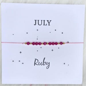 May include: A delicate bracelet featuring a row of small, faceted dark pink beads and tiny gold-toned metallic spacers on a thin light pink cord, displayed on a white card printed with the text JULY and Ruby, surrounded by small black star icons.