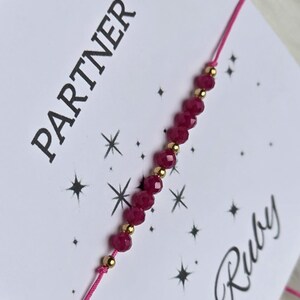 May include: A delicate bracelet featuring alternating small faceted dark pink beads and tiny gold-toned metallic spheres on a thin pink cord. The bracelet is displayed on a white card printed with the word PARTNER and the word Ruby.