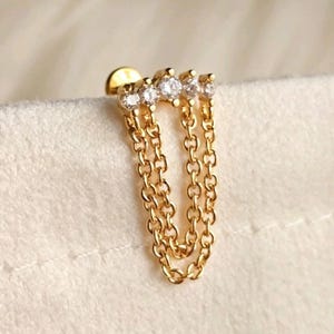 May include: A gold-toned stud earring featuring a horizontal bar set with four small, clear, round-cut stones. Two delicate gold-toned chains hang from the bar, creating a looped, dangling design that connects back to the stud base.