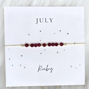 May include: A delicate bracelet featuring a row of small, faceted dark red beads interspersed with tiny gold-coloured metallic spheres on a thin beige cord. The bracelet is displayed on a white card printed with the text JULY and Ruby.