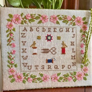 Puede incluir: Un muestrario de punto de cruz con el texto "Stitcher's Rose Garden" y el nombre del diseñador. El muestrario presenta un borde floral de rosas rosas y hojas verdes. El centro incluye el alfabeto, herramientas de costura y un diseño floral.