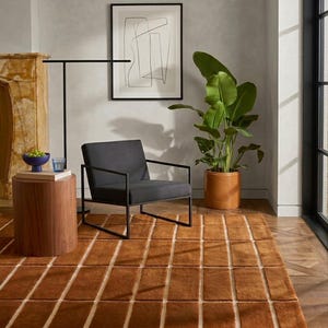 May include: A modern living room featuring a brown rug with a grid pattern, a gray armchair, and a potted plant. A wooden side table and a framed abstract art piece complete the decor. The rug's dimensions are approximately 8 x 10 feet.