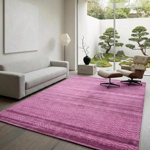 May include: A large, rectangular magenta rug with a textured, geometric pattern. The rug is placed on a wooden floor in a modern living room setting. A grey sofa and a lounge chair are also visible.