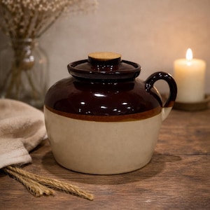 Könnte beinhalten: Zweifarbiger Keramiktopf mit Deckel und Henkel. Der Topf ist oben braun und unten beige, mit einem Holzdeckel. Eine Kerze und Trockenblumen sind im Hintergrund. Der Topf steht auf einer Holzoberfläche.