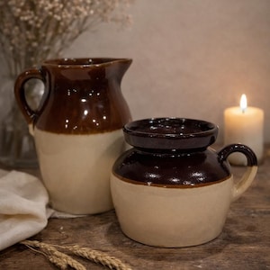 May include: Two-tone ceramic pitchers and a small jar with handles. The upper portion of each piece is a dark brown, while the lower portion is a cream color. The pitcher on the left has a handle and a spout. The jar on the right has a lid.