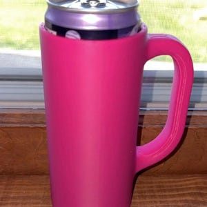 May include: A bright pink can cooler with a handle, holding a purple can. The cooler is cylindrical with a handle on the side. The can is partially visible, with a silver top and a purple body.