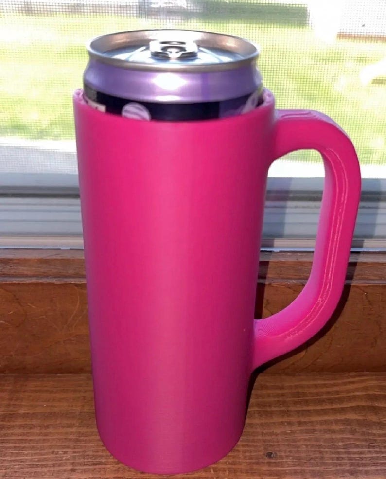 May include: A bright pink can cooler with a handle, holding a purple can. The cooler is cylindrical with a handle on the side. The can is partially visible, with a silver top and a purple body.