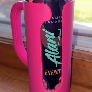 May include: A pink can cooler with a handle holds a can of Alani Nu Energy drink. The can has a dark background with the words "Alani" in teal and "Energy" in white. The cooler is made of plastic.