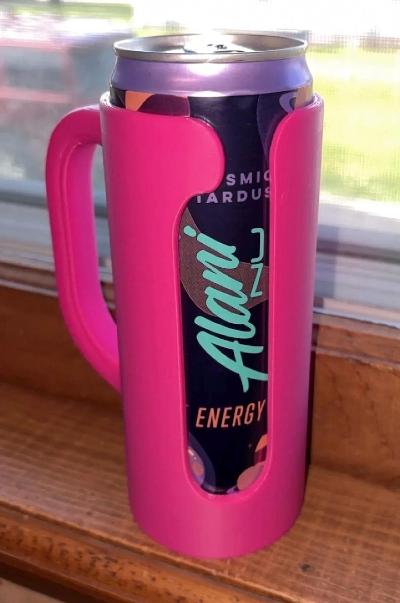 May include: A pink can cooler with a handle holds a can of Alani Nu Energy drink. The can has a dark background with the words "Alani" in teal and "Energy" in white. The cooler is made of plastic.
