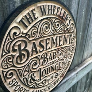 May include: A circular wooden sign with the words "The Wheeler's Basement Bar & Lounge" carved into it. The sign has a dark brown border and a light brown interior with decorative scrollwork. The words "Sports, Games & Entertainment" are also carved into the sign.