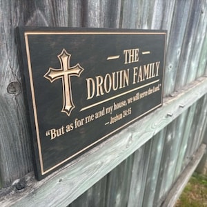 Può includere: Un cartello rettangolare in legno con la scritta "THE DROUIN FAMILY" e un simbolo a croce. Il cartello include anche la citazione "But as for me and my house, we will serve the lord" da Giosuè 24:15. Il cartello è nero con scritte dorate.