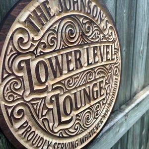 Peut inclure: Panneau rond en bois avec l'inscription gravée "THE JOHNSON'S LOWER LEVEL LOUNGE". Le panneau a un bord marron foncé et un intérieur en bois plus clair avec des volutes décoratives. Le texte "PROUDLY SERVING WHATEVER YOU BROUGHT" est également gravé.