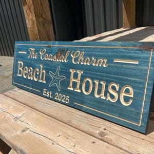 May include: A rectangular blue wooden sign with carved lettering that reads The Coastal Charm Beach House Est. 2025. The sign features a carved starfish graphic centered between the words Beach and House, with horizontal lines above and below.
